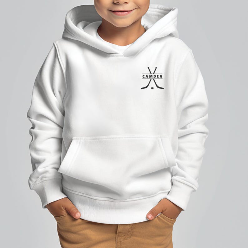 May include: A white hooded sweatshirt featuring a black graphic on the upper left chest. The graphic consists of two crossed hockey sticks with the word CAMDEN printed in black capital letters inside a horizontal rectangular box between them.
