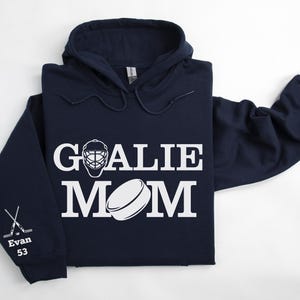 May include: Navy blue hoodie with the words "GOALIE MOM" in white block letters, along with a goalie mask and hockey puck graphic. The left sleeve has crossed hockey sticks and the name "Evan" with the number "53".