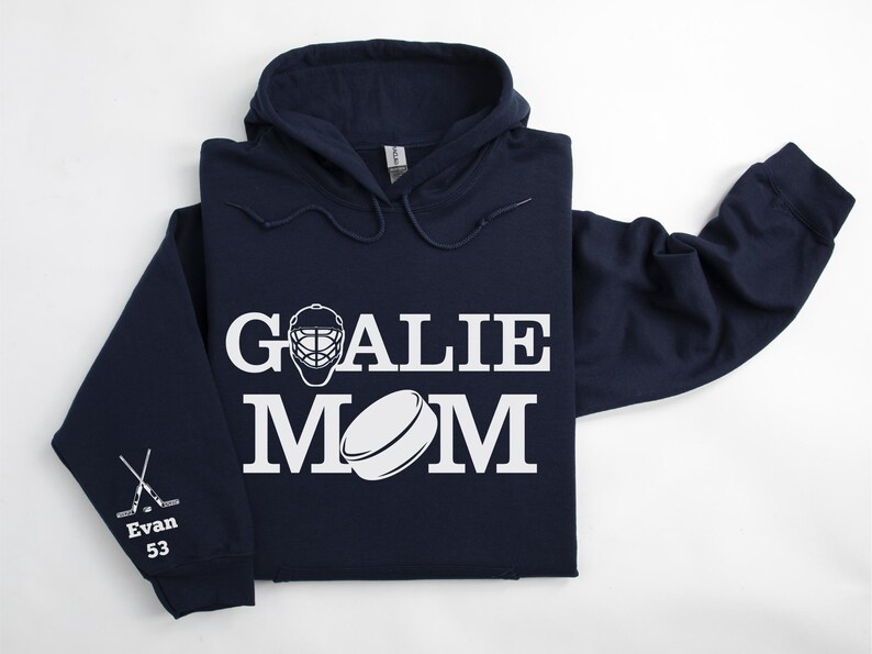 May include: Navy blue hoodie with the words "GOALIE MOM" in white block letters, along with a goalie mask and hockey puck graphic. The left sleeve has crossed hockey sticks and the name "Evan" with the number "53".