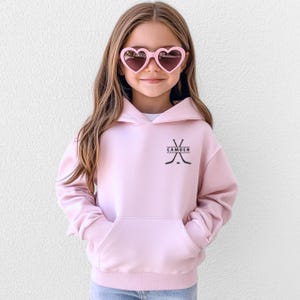 May include: A light pink hooded sweatshirt featuring a black graphic of two crossed hockey sticks and a hockey puck with the name CAMDEN printed in the centre. The garment includes a front kangaroo pocket and long sleeves with ribbed cuffs.
