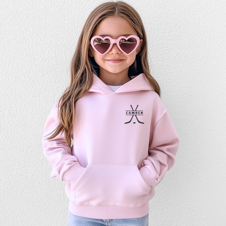 May include: A light pink hooded sweatshirt featuring a black graphic of two crossed hockey sticks and a hockey puck with the name CAMDEN printed in the centre. The garment includes a front kangaroo pocket and long sleeves with ribbed cuffs.