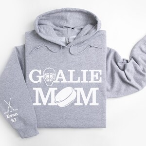 May include: A light gray hoodie with the words "GOALIE MOM" in white, along with a hockey goalie mask and puck graphic. The left sleeve has crossed hockey sticks, the name "Evan", and the number "53".