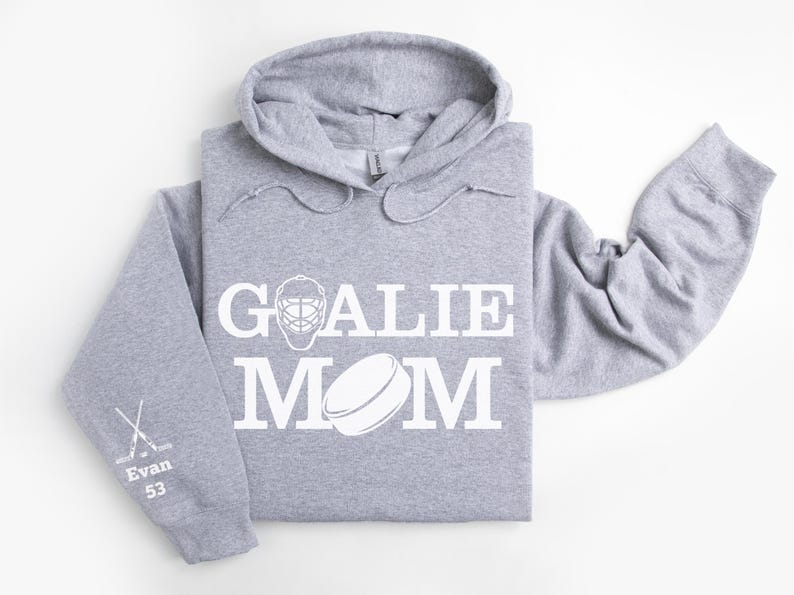 May include: A light gray hoodie with the words "GOALIE MOM" in white, along with a hockey goalie mask and puck graphic. The left sleeve has crossed hockey sticks, the name "Evan", and the number "53".