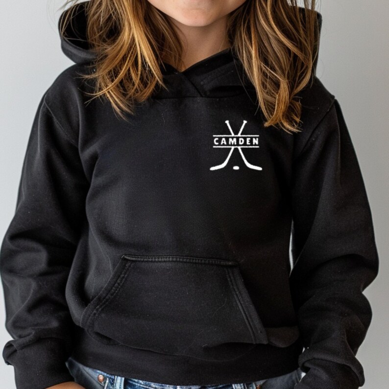 May include: A black hooded sweatshirt featuring a white graphic on the upper left chest. The graphic displays two crossed hockey sticks, a hockey puck, and the text CAMDEN centred within a horizontal rectangular frame between the sticks.