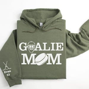 May include: Olive green hoodie with the words "GOALIE MOM" in white block letters, along with a hockey mask and puck graphic. The left sleeve has crossed hockey sticks and the name "Evan 53".