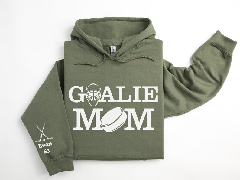 May include: Olive green hoodie with the words "GOALIE MOM" in white block letters, along with a hockey mask and puck graphic. The left sleeve has crossed hockey sticks and the name "Evan 53".