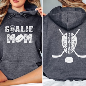 K&ouml;nnte beinhalten: Dunkelgrauer Hoodie mit wei&szlig;em, abgenutztem Text "GOALIE MOM" und einer Hockeypuck-Grafik. Auf der R&uuml;ckseite befinden sich eine Hockey-Torwartmaske, gekreuzte Hockeyschl&auml;ger, ein Puck und der Name "ISAIAH".