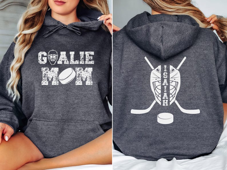 K&ouml;nnte beinhalten: Dunkelgrauer Hoodie mit wei&szlig;em, abgenutztem Text "GOALIE MOM" und einer Hockeypuck-Grafik. Auf der R&uuml;ckseite befinden sich eine Hockey-Torwartmaske, gekreuzte Hockeyschl&auml;ger, ein Puck und der Name "ISAIAH".