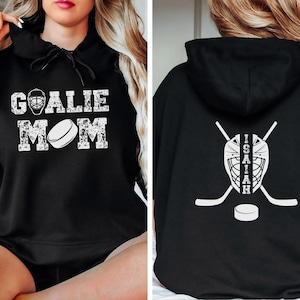 K&ouml;nnte beinhalten: Schwarzer Kapuzenpullover mit dem Text "GOALIE MOM" in verblichener wei&szlig;er Schrift auf der Vorderseite. Auf der R&uuml;ckseite befinden sich eine Hockey-Torwartmaske, gekreuzte Hockeyschl&auml;ger, ein Hockeypuck und der Name "ISAIAH" in Wei&szlig;.