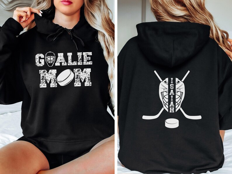 K&ouml;nnte beinhalten: Schwarzer Kapuzenpullover mit dem Text "GOALIE MOM" in verblichener wei&szlig;er Schrift auf der Vorderseite. Auf der R&uuml;ckseite befinden sich eine Hockey-Torwartmaske, gekreuzte Hockeyschl&auml;ger, ein Hockeypuck und der Name "ISAIAH" in Wei&szlig;.
