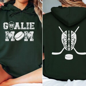 K&ouml;nnte beinhalten: Dunkelgr&uuml;ner Kapuzenpullover mit wei&szlig;em, verwaschenem Text und Grafiken. Auf der Vorderseite steht "GOALIE MOM" mit einer Hockeymaske und einem Puck. Auf der R&uuml;ckseite befinden sich eine Hockeymaske, gekreuzte Hockeyschl&auml;ger, ein Puck und der Name "ISAIAH".
