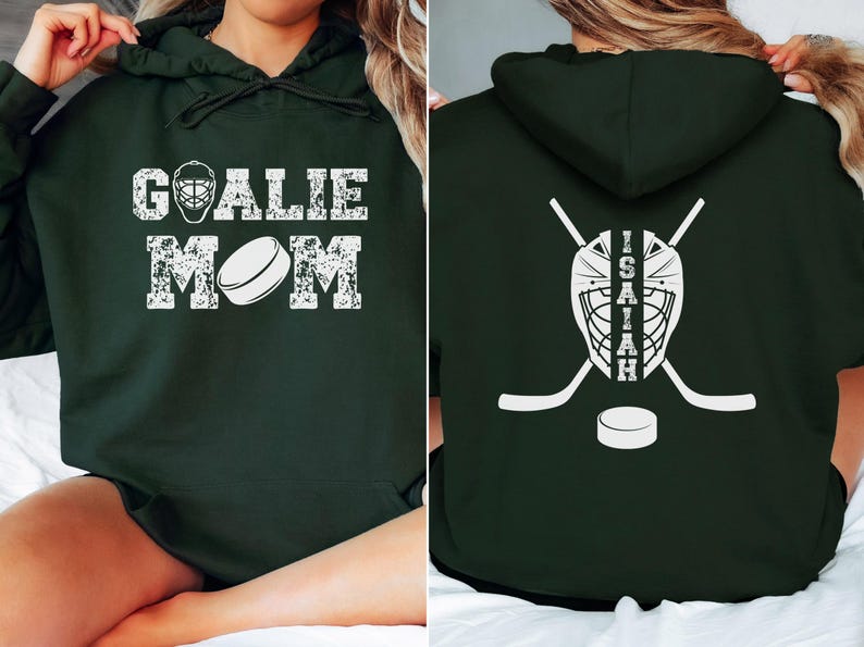 K&ouml;nnte beinhalten: Dunkelgr&uuml;ner Kapuzenpullover mit wei&szlig;em, verwaschenem Text und Grafiken. Auf der Vorderseite steht "GOALIE MOM" mit einer Hockeymaske und einem Puck. Auf der R&uuml;ckseite befinden sich eine Hockeymaske, gekreuzte Hockeyschl&auml;ger, ein Puck und der Name "ISAIAH".