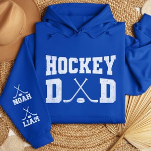 Peut inclure: Un sweat &agrave; capuche bleu roi avec les mots "HOCKEY DAD" en lettres blanches. Les manches portent les noms "NOAH" et "LIAM" avec des graphiques de crosses de hockey. Un chapeau marron est en arri&egrave;re-plan.