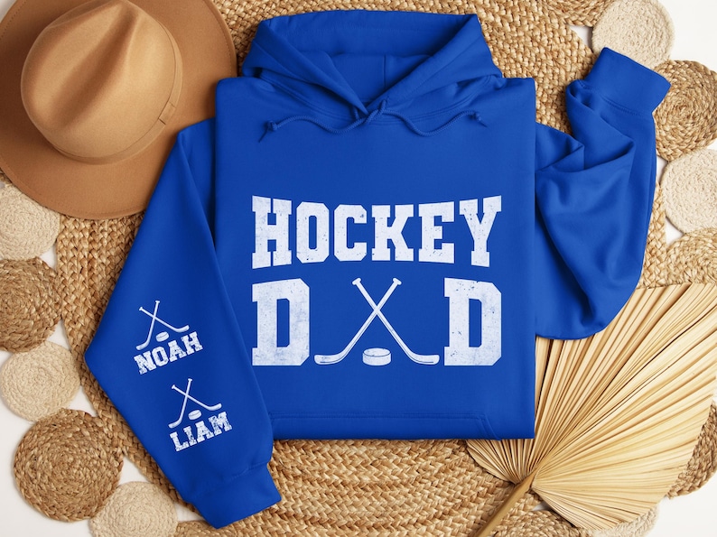 Peut inclure: Un sweat &agrave; capuche bleu roi avec les mots "HOCKEY DAD" en lettres blanches. Les manches portent les noms "NOAH" et "LIAM" avec des graphiques de crosses de hockey. Un chapeau marron est en arri&egrave;re-plan.