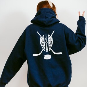 K&ouml;nnte beinhalten: Marineblauer Kapuzenpullover mit wei&szlig;em Hockey-Motiv auf dem R&uuml;cken. Das Design zeigt eine Torwartmaske, gekreuzte Hockeyschl&auml;ger, einen Puck und den Namen "Isaiah". Der Hoodie hat eine Kapuze und lange &Auml;rmel.