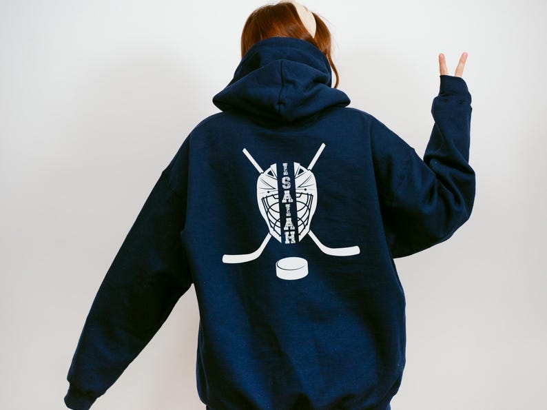 K&ouml;nnte beinhalten: Marineblauer Kapuzenpullover mit wei&szlig;em Hockey-Motiv auf dem R&uuml;cken. Das Design zeigt eine Torwartmaske, gekreuzte Hockeyschl&auml;ger, einen Puck und den Namen "Isaiah". Der Hoodie hat eine Kapuze und lange &Auml;rmel.