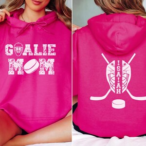 K&ouml;nnte beinhalten: Ein magentafarbener Kapuzenpullover mit wei&szlig;em Text und Grafiken. Auf der Vorderseite steht "GOALIE MOM" mit einer Hockeymaske und einer Puckgrafik. Auf der R&uuml;ckseite befinden sich eine Hockeymaske, gekreuzte Hockeyschl&auml;ger, ein Puck und der Name "ISAIAH".