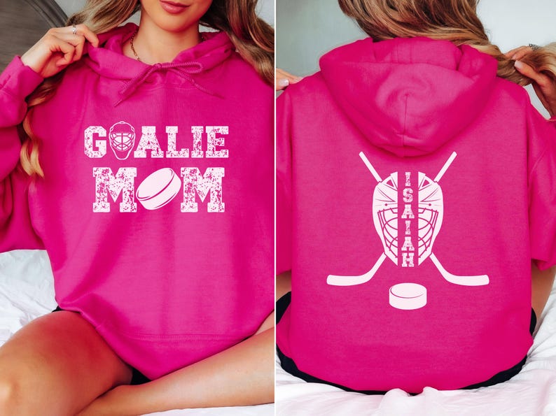 K&ouml;nnte beinhalten: Ein magentafarbener Kapuzenpullover mit wei&szlig;em Text und Grafiken. Auf der Vorderseite steht "GOALIE MOM" mit einer Hockeymaske und einer Puckgrafik. Auf der R&uuml;ckseite befinden sich eine Hockeymaske, gekreuzte Hockeyschl&auml;ger, ein Puck und der Name "ISAIAH".