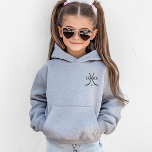 May include: A heather grey hooded sweatshirt featuring a front kangaroo pocket and a black graphic on the chest depicting two crossed hockey sticks with the text CAMDEN centred between them. The hoodie has long sleeves and a hood.