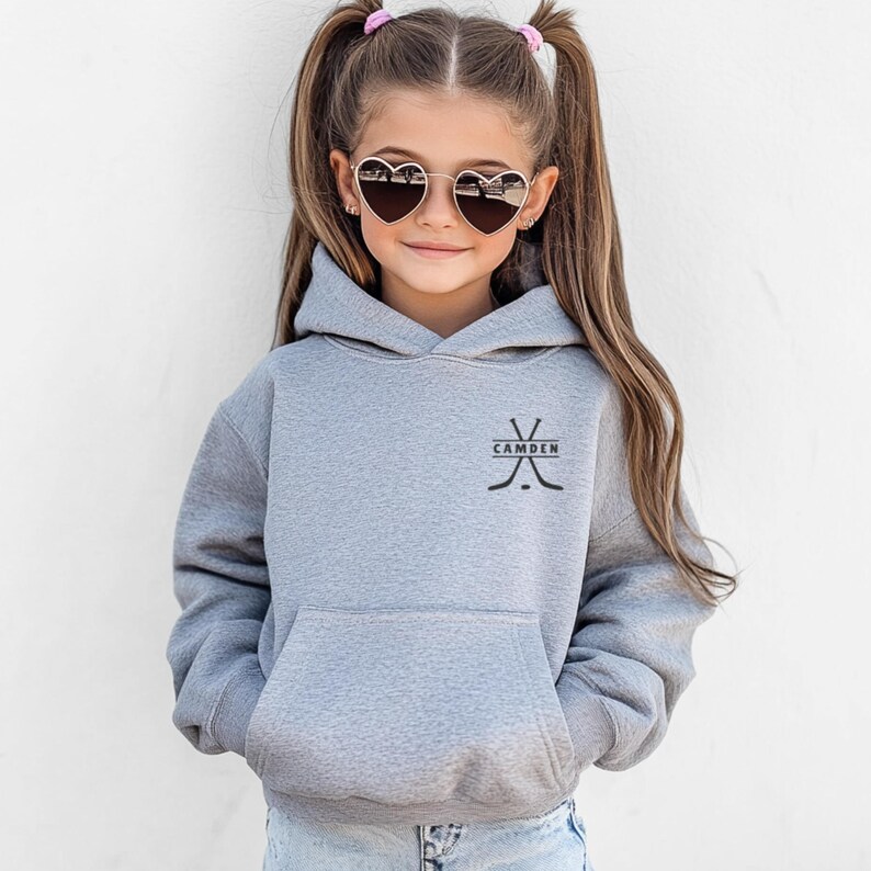May include: A heather grey hooded sweatshirt featuring a front kangaroo pocket and a black graphic on the chest depicting two crossed hockey sticks with the text CAMDEN centred between them. The hoodie has long sleeves and a hood.