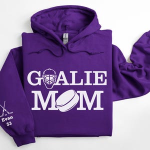May include: A purple hoodie with the words "GOALIE MOM" in white, along with a hockey goalie mask and a hockey puck graphic. The left sleeve has crossed hockey sticks, the name "Evan" and the number "53".