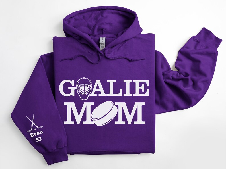 May include: A purple hoodie with the words "GOALIE MOM" in white, along with a hockey goalie mask and a hockey puck graphic. The left sleeve has crossed hockey sticks, the name "Evan" and the number "53".