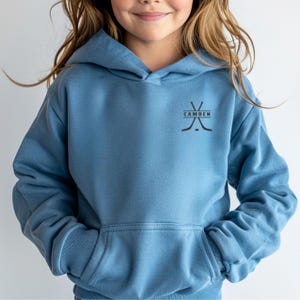 May include: A dusty blue hooded sweatshirt featuring a front kangaroo pocket and long sleeves. On the upper left chest, there is a black graphic design consisting of two crossed hockey sticks with the word CAMDEN printed horizontally between them.