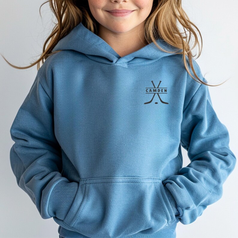 May include: A dusty blue hooded sweatshirt featuring a front kangaroo pocket and long sleeves. On the upper left chest, there is a black graphic design consisting of two crossed hockey sticks with the word CAMDEN printed horizontally between them.