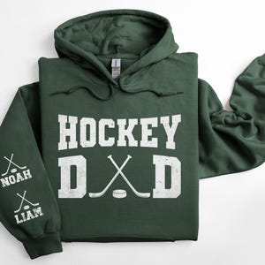 Peut inclure: Un sweat &agrave; capuche vert fonc&eacute; avec le texte "HOCKEY DAD" en blanc. Le design comprend des crosses de hockey crois&eacute;es et un palet. La manche gauche affiche les noms "NOAH" et "LIAM" avec des graphiques de crosses.