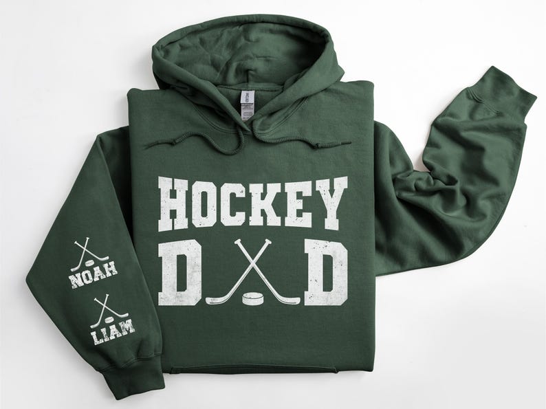 Peut inclure: Un sweat &agrave; capuche vert fonc&eacute; avec le texte "HOCKEY DAD" en blanc. Le design comprend des crosses de hockey crois&eacute;es et un palet. La manche gauche affiche les noms "NOAH" et "LIAM" avec des graphiques de crosses.