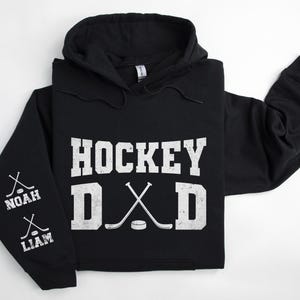 May include: Black hoodie with the text "HOCKEY DAD" in white, along with crossed hockey sticks and a puck graphic. The left sleeve features the names "NOAH" and "LIAM" with hockey stick graphics. A casual sports-themed apparel item.