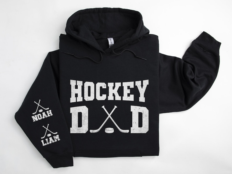 Peut inclure: Sweat &agrave; capuche noir avec l'inscription blanche "HOCKEY DAD", des crosses de hockey crois&eacute;es et un palet. La manche gauche affiche les noms "NOAH" et "LIAM" avec des graphiques de crosses. V&ecirc;tement d&eacute;contract&eacute; sur le th&egrave;me du sport.