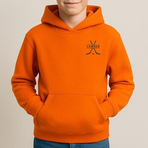 May include: A bright orange hooded sweatshirt featuring a front kangaroo pocket and a black graphic on the upper left chest. The graphic displays two crossed hockey sticks with the word CAMDEN printed in black capital letters between them.