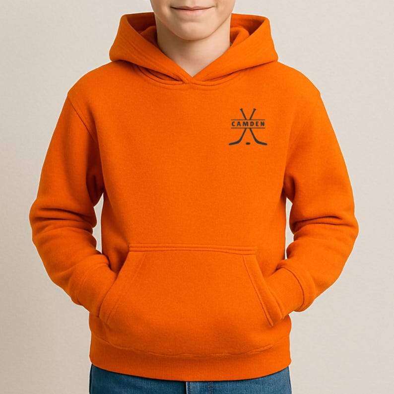 May include: A bright orange hooded sweatshirt featuring a front kangaroo pocket and a black graphic on the upper left chest. The graphic displays two crossed hockey sticks with the word CAMDEN printed in black capital letters between them.