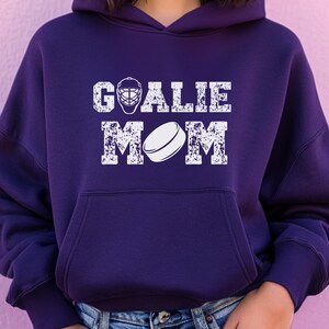 K&ouml;nnte beinhalten: Dunkelvioletter Hoodie mit wei&szlig;em Aufdruck &bdquo;GOALIE MOM&ldquo;. Der Aufdruck zeigt eine Hockey-Torwartmaske und einen Hockeypuck. Der Hoodie hat eine Vordertasche und eine Kapuze.