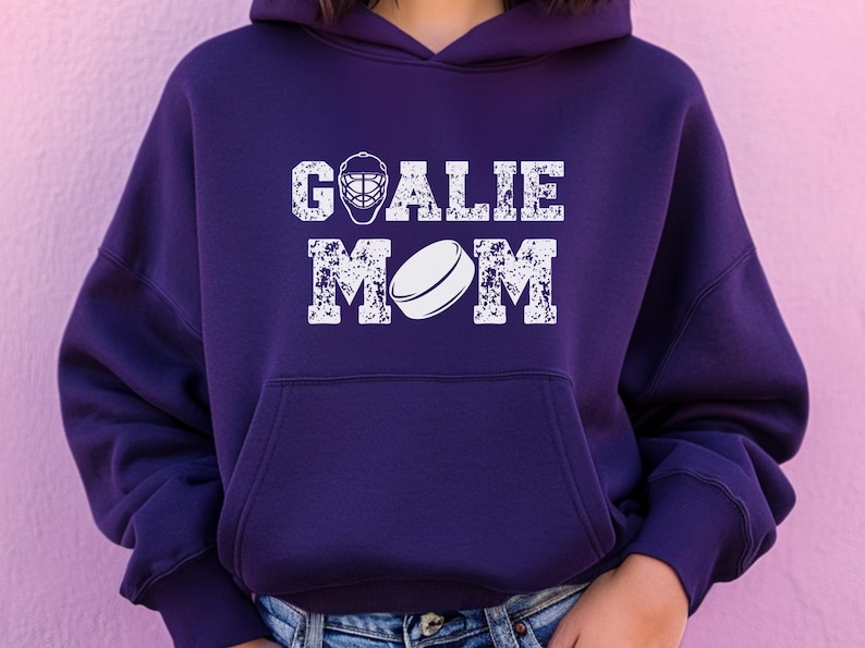 K&ouml;nnte beinhalten: Dunkelvioletter Hoodie mit wei&szlig;em Aufdruck &bdquo;GOALIE MOM&ldquo;. Der Aufdruck zeigt eine Hockey-Torwartmaske und einen Hockeypuck. Der Hoodie hat eine Vordertasche und eine Kapuze.