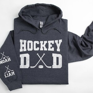 Peut inclure: Un sweat &agrave; capuche gris fonc&eacute; avec l'inscription blanche "HOCKEY DAD". Le design comprend des crosses de hockey crois&eacute;es et une rondelle. La manche affiche les noms "NOAH" et "LIAM" avec des graphiques de crosses.