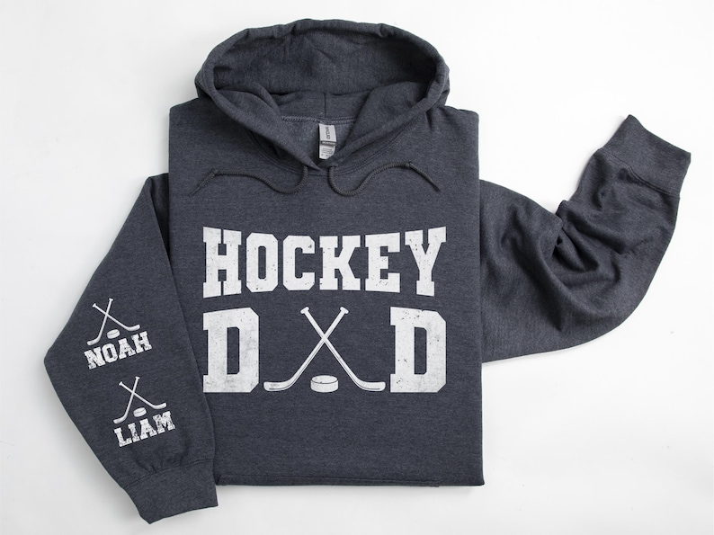 Peut inclure: Un sweat &agrave; capuche gris fonc&eacute; avec l'inscription blanche "HOCKEY DAD". Le design comprend des crosses de hockey crois&eacute;es et une rondelle. La manche affiche les noms "NOAH" et "LIAM" avec des graphiques de crosses.