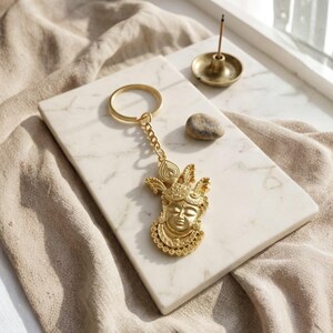 May include: A gold-toned metal keychain featuring a detailed, embossed face of a deity with an ornate headdress and circular necklace. The charm is attached to a standard circular split ring by a short chain, showcasing intricate relief work.