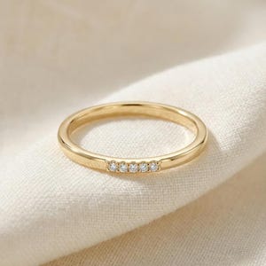 Puede incluir: Un anillo dorado con cinco pequeñas piedras preciosas transparentes dispuestas en fila. El anillo tiene una banda lisa y pulida. El anillo se presenta sobre una tela blanca. El diseño del anillo es sencillo y elegante.
