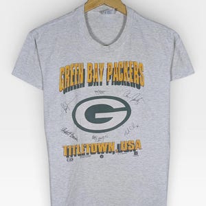Puede incluir: Una camiseta gris con la inscripción "GREEN BAY PACKERS" en dorado y verde, el logotipo del equipo y "TITLETOWN, USA". La camiseta tiene mangas cortas y cuello redondo. Varias firmas son visibles en la camiseta.