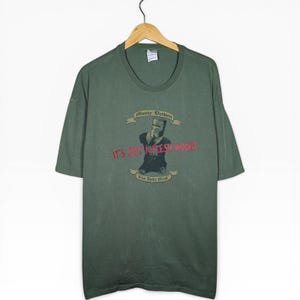 May include: A green t-shirt featuring a graphic from Monty Python and the Holy Grail. The graphic shows a knight with the text "It's Just a Flesh Wound". The shirt is suspended from a wooden hanger.