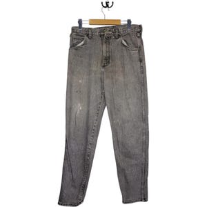 May include: Grey denim jeans hanging on a wooden hanger. The jeans feature a classic five-pocket design, a button and zip closure, and show signs of wear with some light discolouration.
