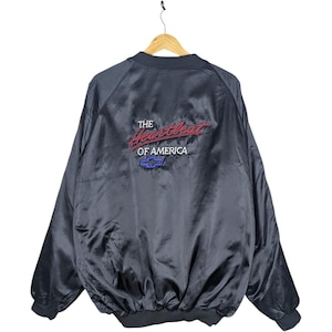 Puede incluir: Una chaqueta bomber de satén azul marino oscuro con mangas largas y cuello acanalado. La parte posterior muestra el texto bordado The Heartbeat OF AMERICA en rojo y blanco, acompañado por un logotipo azul de Chevrolet en forma de pajarita.