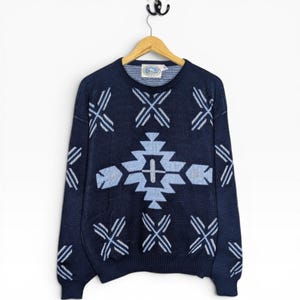 May include: A navy blue sweater featuring a geometric pattern in light blue and white. The sweater is suspended on a wooden hanger. The design includes cross and arrow-like shapes. The background is white.