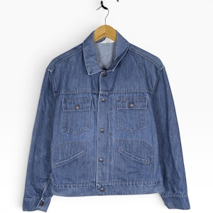 May include: A vintage blue denim jacket with a classic design. The jacket features a collared neckline, button-down front, two chest pockets, and two lower pockets. It is hanging on a wooden hanger against a white background.