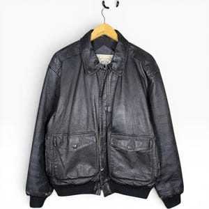 Könnte beinhalten: Eine schwarze Lederjacke mit klassischem Kragen, durchgehendem Frontreißverschluss und zwei großen Vordertaschen mit Druckknöpfen. Die Jacke verfügt über gerippte Bündchen und einen gerippten Saum sowie ein sichtbares Innenetikett am Halsausschnitt.