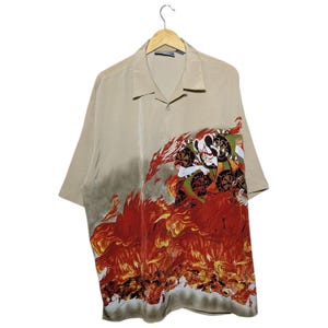 Puede incluir: Una camisa beige de manga corta con un gráfico llamativo. El diseño presenta una ilustración de inspiración japonesa de una figura en medio de llamas en tonos de rojo, naranja y blanco. La camisa está colgada de una percha de madera.