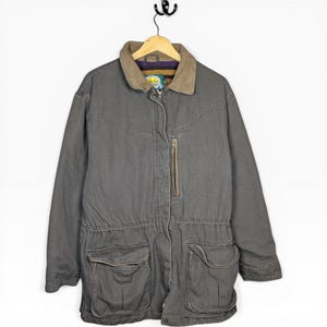 May include: A gray Cabela's jacket with a brown corduroy collar and multiple pockets. The jacket has a zipper closure and a drawstring waist. The interior lining is visible at the collar. The jacket is hanging on a wooden hanger.