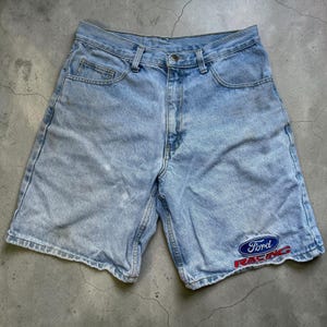 Puede incluir: Pantalones cortos de mezclilla azul claro con un diseño clásico de cinco bolsillos, cierre de botón y cremallera. La pierna inferior izquierda muestra un parche bordado con el logotipo de Ford Racing en hilo azul, blanco y rojo.